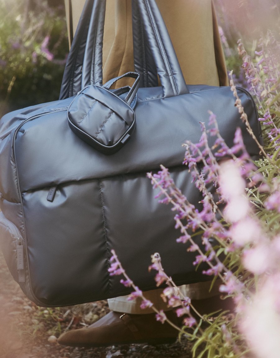 Calpak spot — a navy quilted weekender carried along a garden path